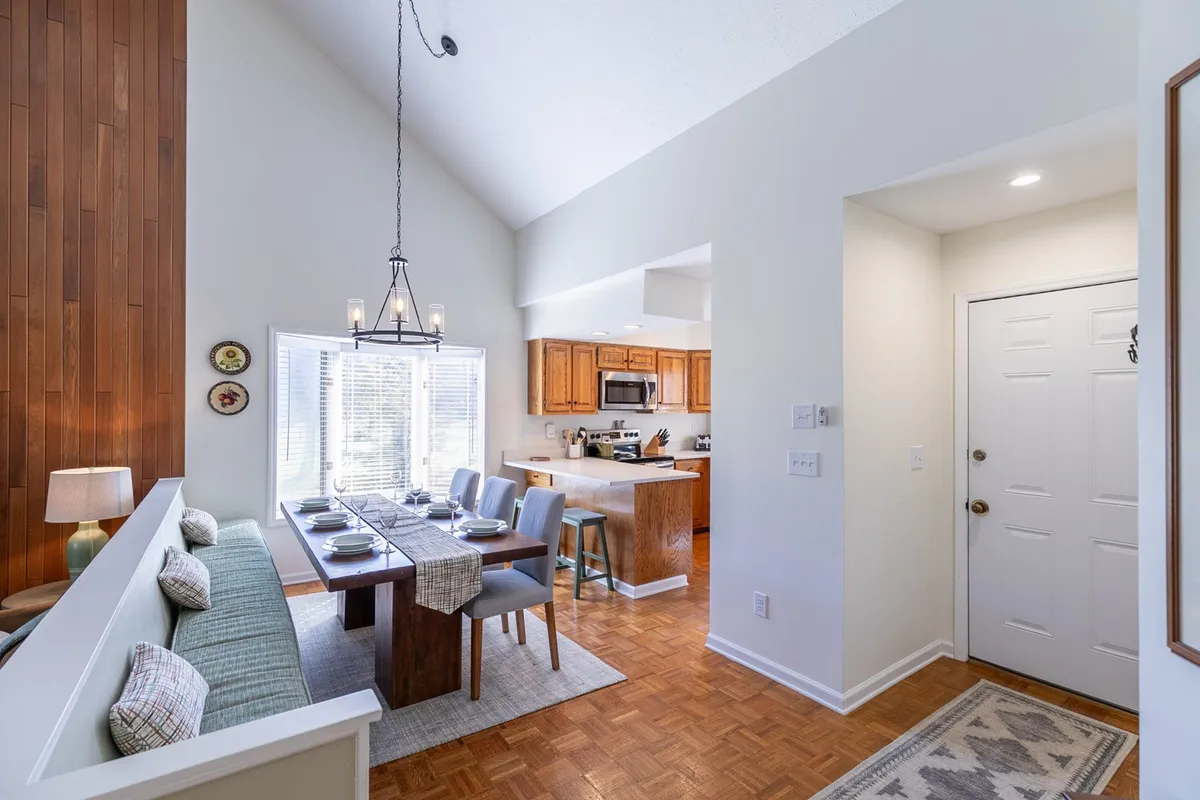 A cozy dining area is pictured, with a dining table and chairs set against a wooden wall. The kitchen is visible in the background, creating a sense of open, connected living. The scene is inviting, perfect for family meals or casual gatherings.