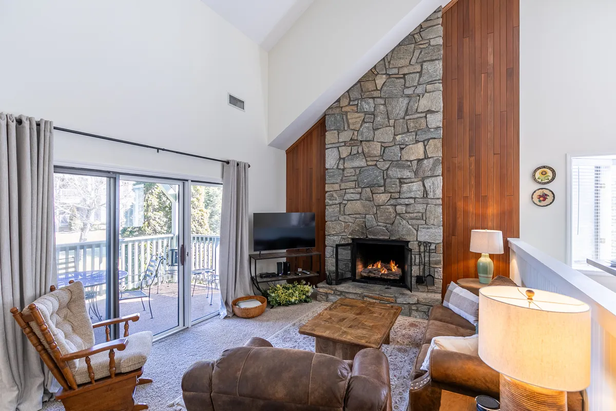 The hero image showcases a warm, inviting living room with a beautiful stone gas fireplace as the centerpiece, surrounded by plush seating, a rustic wooden coffee table, exposed beams, and large windows that bathe the room in natural light. Decorative elements on the wall and cozy furnishings add to the welcoming mountain retreat atmosphere.