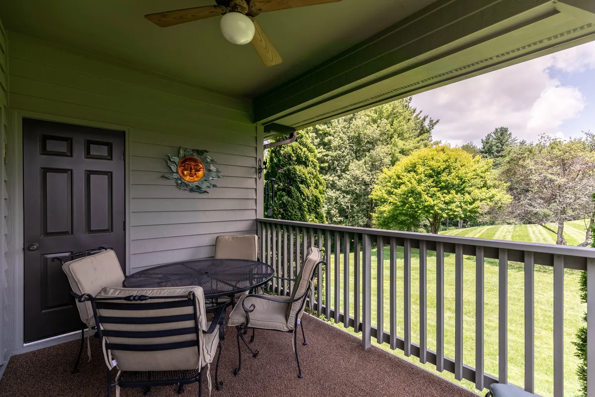 A covered outdoor patio with a round glass table and chairs sits next to a sliding door, surrounded by lush greenery and shaded by mature trees. The inviting patio provides a tranquil setting for outdoor dining or group gatherings, with subtle decorative touches enhancing the upscale ambiance.