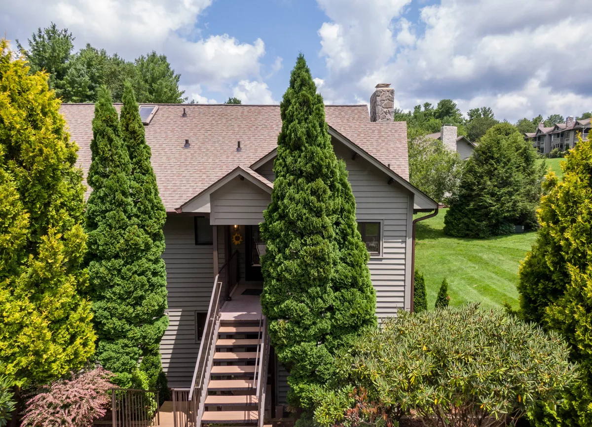 The welcoming exterior of the condo, with entrance steps framed by tall conifer trees and a prominent stone chimney. The setting feels inviting for arrivals and hints at the tranquil, landscaped grounds surrounding the property.