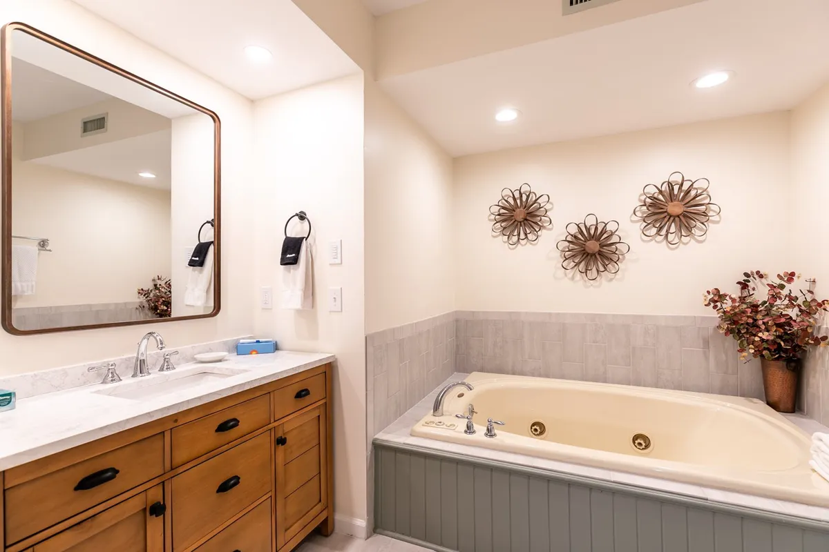 A serene and luxe bathroom setting featuring a soaking bathtub, elegant vanity with a mirror, decorative wall art, and a fresh flower arrangement—evoking spa-like tranquility.