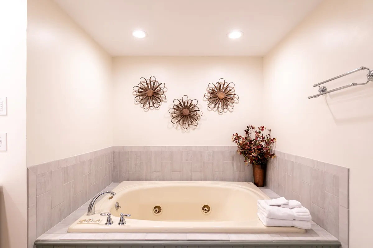 A luxurious bathroom with a large jetted tub, elegant decorative wall art, floral arrangements, and plush towels. The soft lighting and spa-like atmosphere create an inviting space for relaxation and rejuvenation.