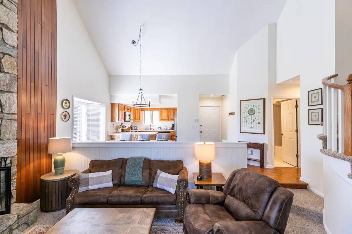 A cozy living area focused around a substantial stone fireplace, with a leather sofa and wooden coffee table providing comfortable seating. The kitchen is visible through a pass-through, reinforcing the open-concept layout. Wood accents and a warm color palette add to the inviting atmosphere.