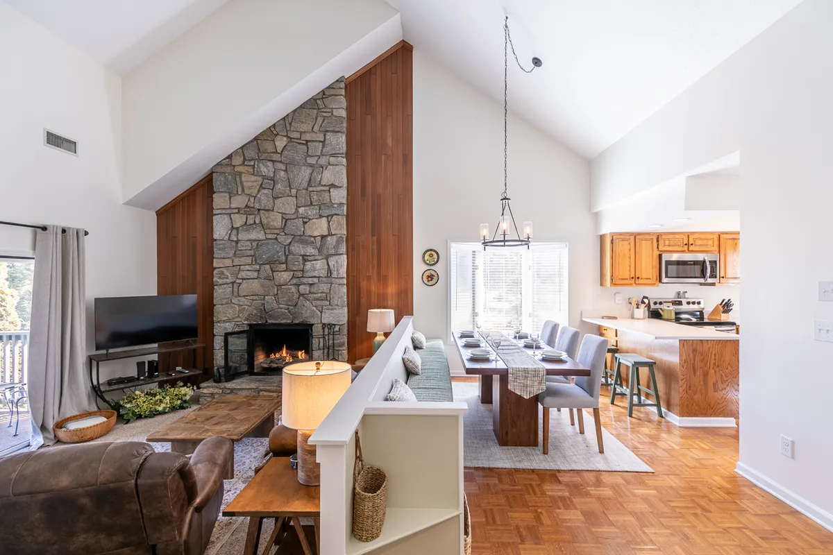 This image presents a spacious open-concept area anchored by a prominent stone fireplace. Plush seating and a cozy living room couch face the fire, while a dining table and kitchen area are seamlessly visible in the background. The overall atmosphere is warm and inviting, and natural light streams in from the windows, enhancing the sense of space.