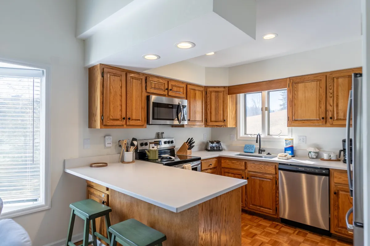 A modern kitchen with a spacious island, stainless steel appliances, wooden cabinetry, bar stools, and ample natural light streaming through a large window—showcasing upscale finishes and a welcoming space for gathering.