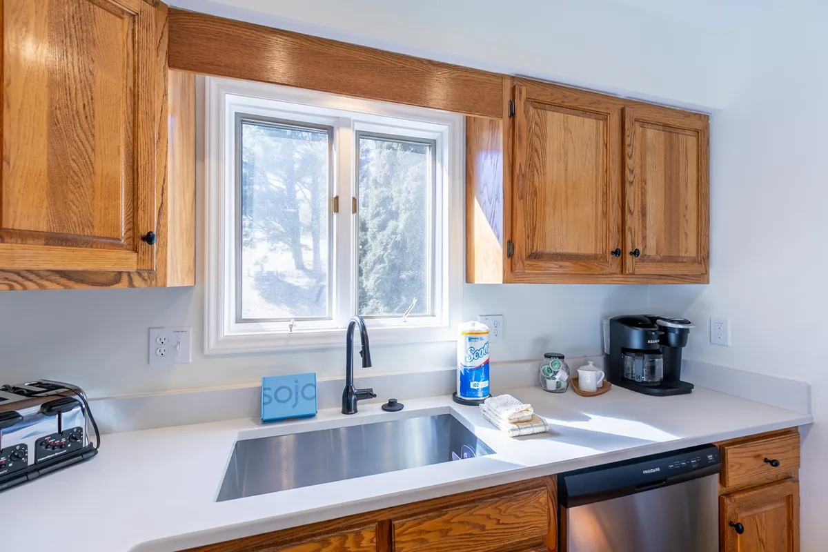 A kitchen scene featuring a window streaming in natural light, a kitchen sink, and a coffee maker on the countertop. The view captures a cozy spot ideal for morning coffee rituals and casual conversation.