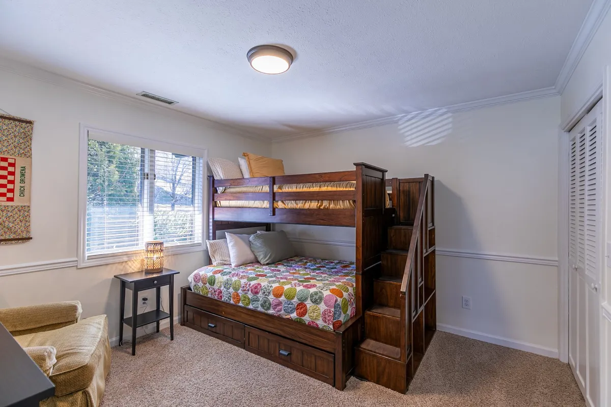 A bright, cheerful bunk room featuring a sturdy twin-over-full bunk bed, a colorful quilt, and a nightstand with a lamp for bedtime reading or nighttime comfort. The playful, welcoming design is especially appealing to younger guests.